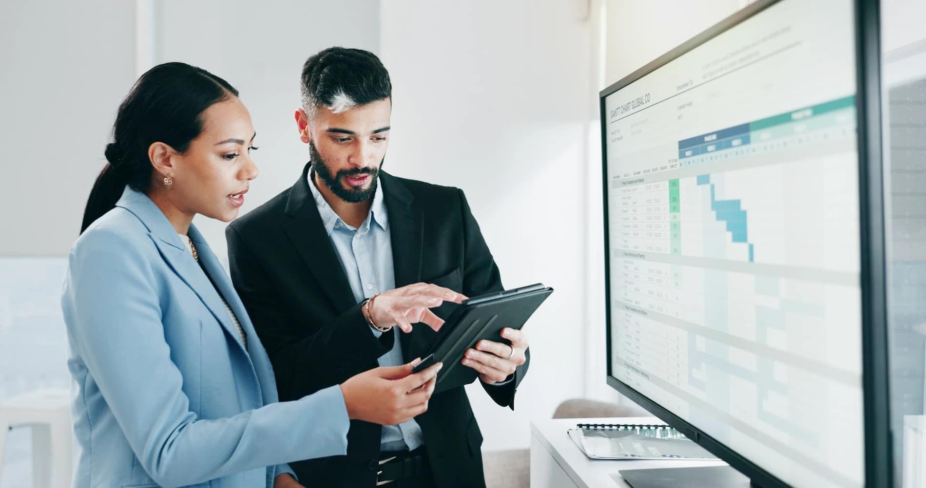 Zwei Geschäftsleute arbeiten gemeinsam an Datenintegrationsstrategien für das UN-Ökosystem und betrachten Analysen auf einem Tablet und einem großen Bildschirm in einem modernen Büro
