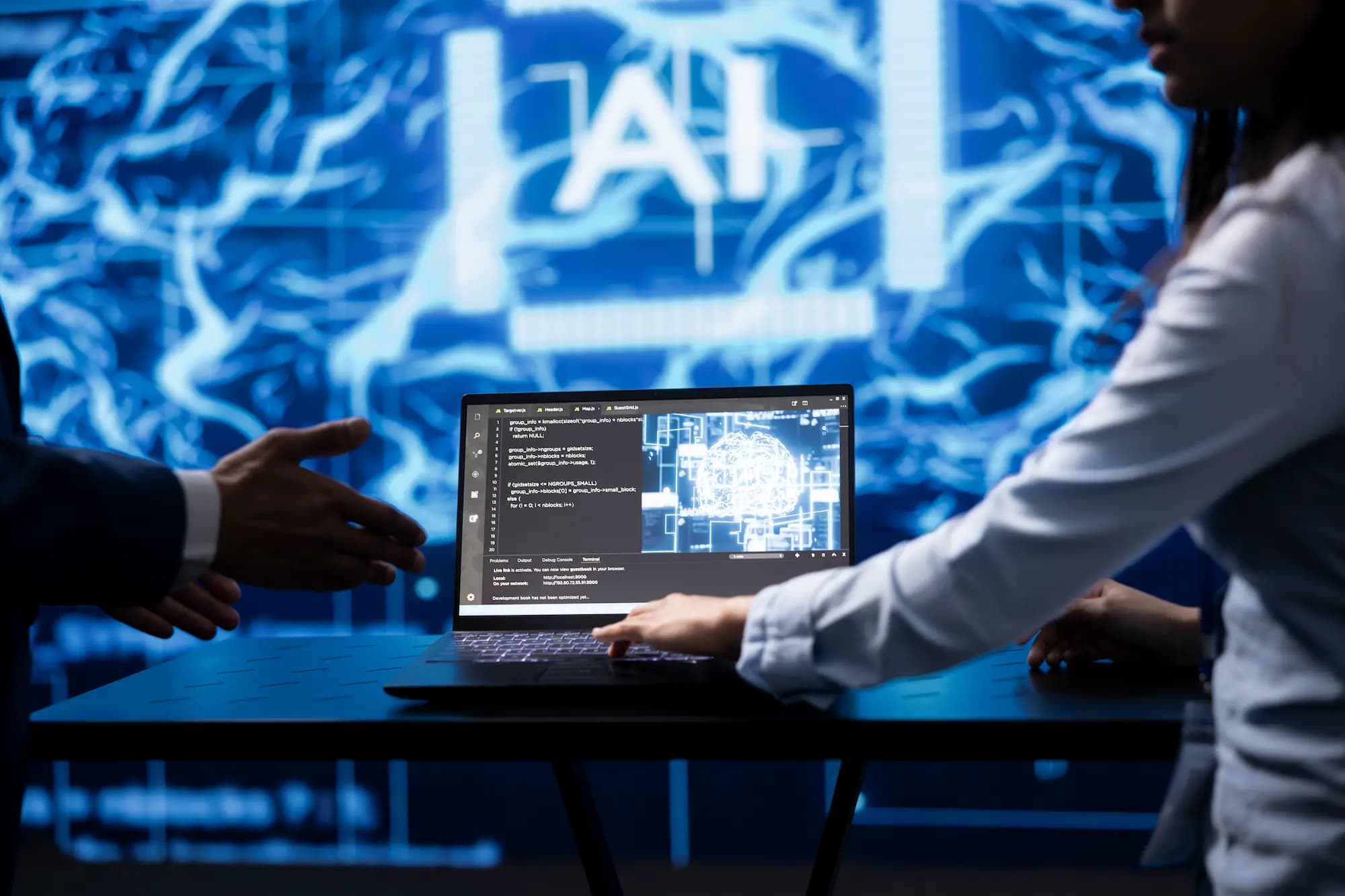 Developers collaborating at a laptop displaying source code and an AI model visualization on a large screen in the background, representing AI-assisted software development and code review.