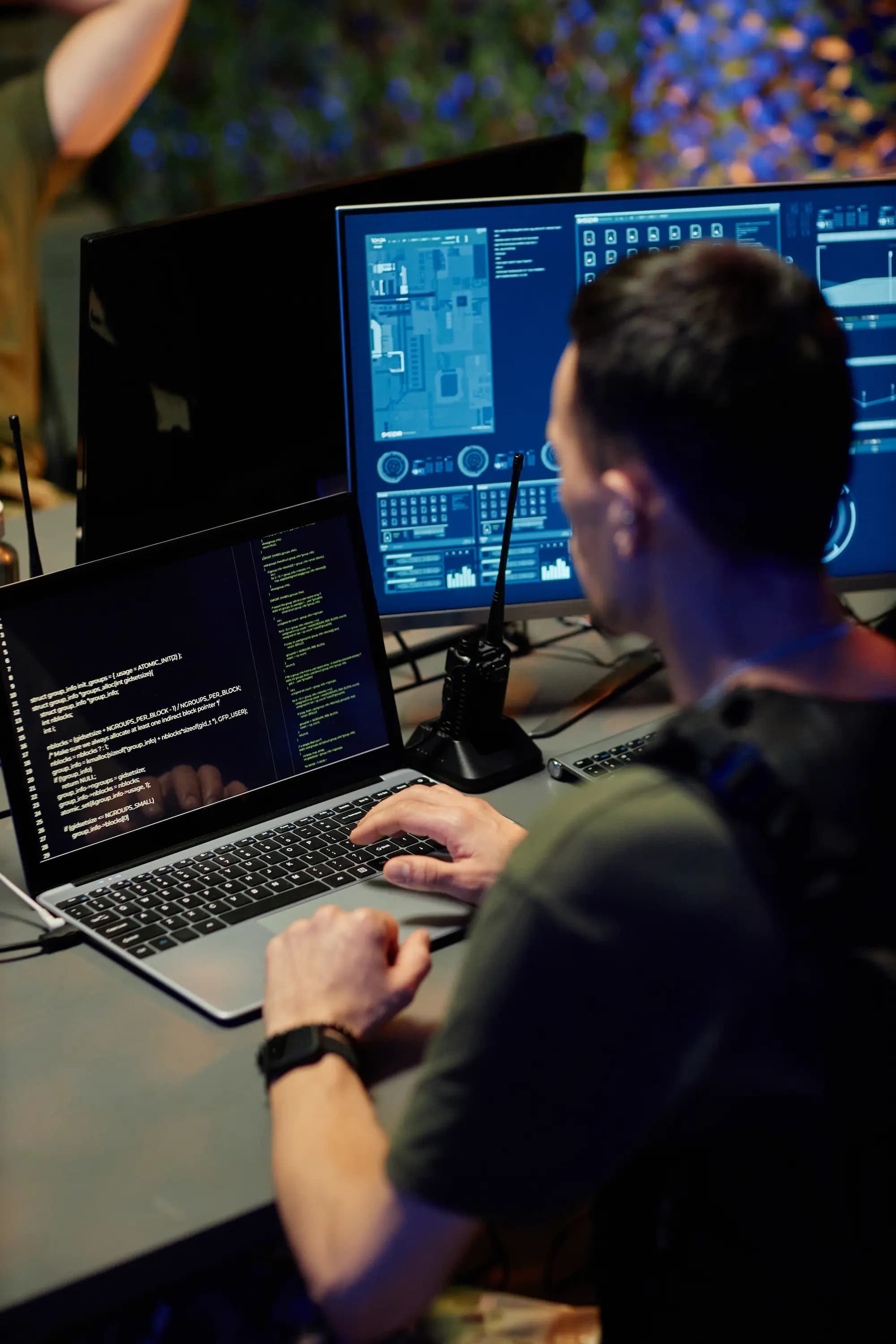 Cybersecurity analyst monitoring code and network data on multiple screens while working on a laptop in a security operations center